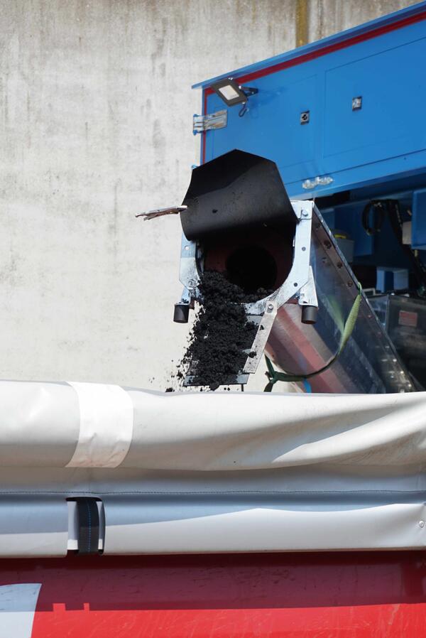 Schwarzer Klärschlamm fällt v on einem Förderband in einen Container